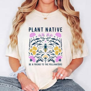 Op de afbeelding: Een crèmekleurig T-shirt met korte mouwen en een grafische print met de tekst PLANT NATIVE en BE A FRIEND TO THE POLLINATORS. Het ontwerp toont een bij in het midden, omringd door donkergroen gebladerte, gele bloemen en roze bloesems.