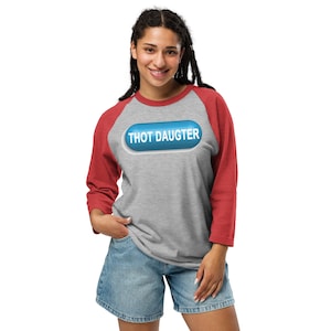 May include: A gray baseball tee with red sleeves. The shirt features a blue pill-shaped graphic with the words "THOT DAUGTER" in white. The person is wearing denim shorts.