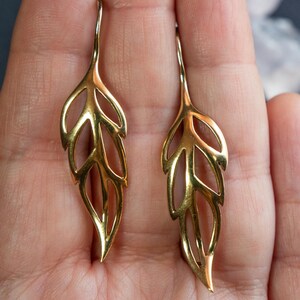 May include: A pair of gold leaf-shaped earrings with a delicate, openwork design. The earrings are held in a hand, showcasing their intricate details.