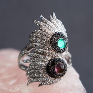 May include: A silver ring with a black and white feather design and two gemstones. The top gemstone is green and the bottom gemstone is purple.