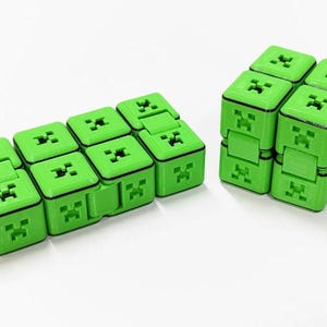Puede incluir: Dos juguetes fidget verdes con forma de bloques de Minecraft. Uno es un cubo y el otro es un prisma rectangular.