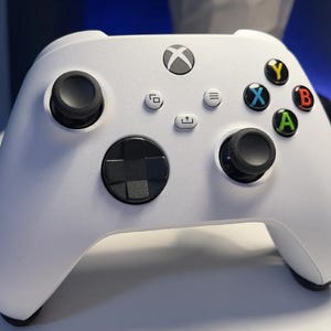 May include: A white Xbox Series X controller with black buttons and a black directional pad. The controller has a white Xbox logo on the front.