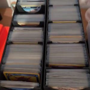 May include: Two black card storage boxes with clear plastic dividers. The boxes are filled with trading cards. The top card in the left box shows a Pokemon card with the name "Claydol" printed on it. The top card in the right box shows a Pokemon card with the name "Wailord" printed on it.