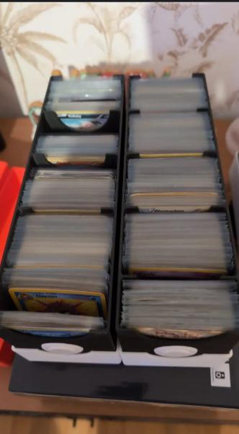May include: Two black card storage boxes with clear plastic dividers. The boxes are filled with trading cards. The top card in the left box shows a Pokemon card with the name "Claydol" printed on it. The top card in the right box shows a Pokemon card with the name "Wailord" printed on it.