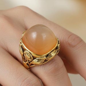 May include: A gold-colored ring featuring a large, peach-colored gemstone. The ring has a detailed floral and leaf design around the stone. The ring is on a person's finger.