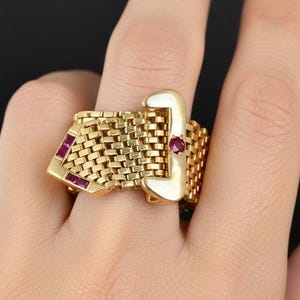 May include: A gold-toned ring with a buckle design. The ring features a woven band and a rectangular buckle with a red gemstone. Additional red gemstones are set into the ring's design. The ring is on a finger.