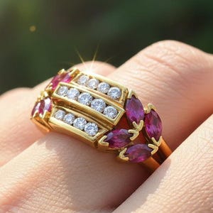 Pode incluir: Um anel de ouro com três fileiras de pequenas pedras preciosas redondas e transparentes e várias pedras preciosas vermelhas profundas em forma de marquesa. O anel se destaca em um fundo verde desfocado, destacando o brilho e o design da joia.