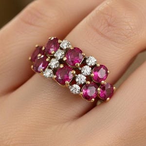 May include: A gold ring featuring a cluster of oval-cut, deep red gemstones and small, round, white gemstones. The ring is worn on a person's finger, showcasing the intricate design and vibrant colors of the jewelry.