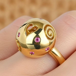 May include: A gold ring featuring a spherical design with heart and swirl cutouts. The sphere is adorned with small pink gemstones. The ring is a bright, polished gold color and is worn on a finger.