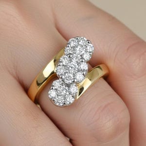 May include: A gold ring with three clusters of small, round, clear gemstones. The ring's band curves around the finger, and the gemstones are set in a cluster design. The ring is on a person's finger.