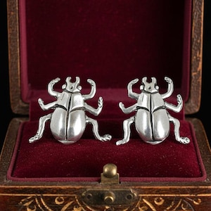 Puede incluir: Un par de gemelos de metal plateado con forma de escarabajo, con patas detalladas y cuerpos segmentados, presentados dentro de una pequeña caja de madera rectangular con un forro de terciopelo rojo oscuro y un cierre de metal.
