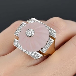 May include: A gold and silver ring featuring a light pink, flower-shaped stone with a central diamond. The ring is set with additional diamonds around the flower and on the geometric frame.
