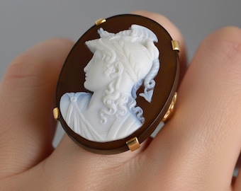 May include: An oval ring featuring a dark brown stone base with a carved white relief of a profile figure wearing a helmet. The cameo is set in a gold-toned metal frame with four small prongs securing the stone to the band on the finger.