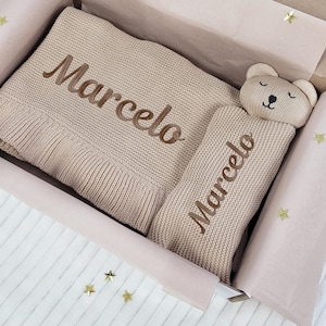 Peut inclure: Un coffret cadeau pour bébé beige, comprenant une couverture tricotée et un oreiller en forme d'ours, tous deux brodés du nom "Marcelo" en fil marron. La couverture est pliée et l'oreiller est à côté.
