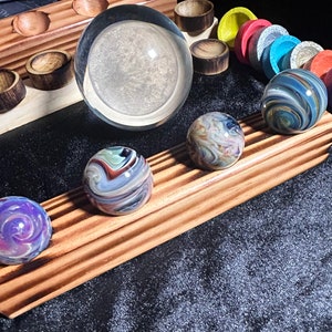May include: Four colorful glass marbles with swirling patterns are displayed on a wooden stand. The marbles are arranged in a row, with a clear glass sphere in the background.