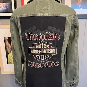 Könnte beinhalten: Olivgrüne Jeansjacke mit schwarzem Rückenteil. Auf dem Panel ist das Harley-Davidson Cycles-Logo und die Slogans "Live to Ride" und "Ride to Live" im Used-Look abgebildet. Die Jacke hat einen Knopfverschluss und einen Kragen.
