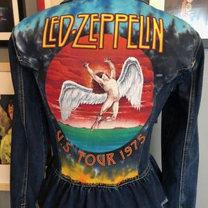 May include: Dark blue denim jacket with a painted Led Zeppelin design on the back. The design features the band's name in yellow, a winged figure, and "U.S. TOUR 1975". The jacket has a ruffled hem.