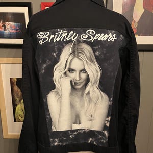 May include: Black denim jacket with a black and white image of Britney Spears on the back. The name "Britney Spears" is written in white script above the image. The jacket has a classic collar and button fastenings.