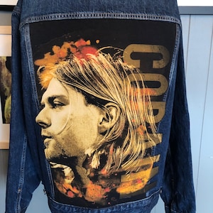 Może przedstawiać: Niebieska dżinsowa kurtka z czarną naszywką z tyłu. Naszywka przedstawia portret Kurta Cobaina z żółtym i pomarańczowym efektem akwarelowym i napisem "Cobain" złotymi literami.