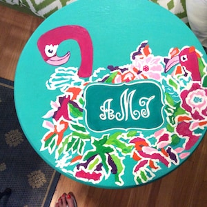 May include: A round, turquoise painted table top with a flamingo and floral design. The flamingo is pink with a white beak and eye. The flowers are pink, purple, orange, and white. The initials "A M J" are in a white frame.
