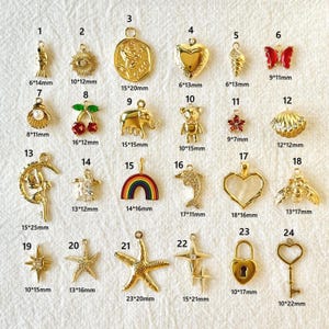 Könnte beinhalten: Eine Sammlung goldfarbener Anhänger in verschiedenen Formen, darunter Fisch, Sonne, Rose, Herz, Muschel, Schmetterling, Perle, Kirschen, Elefant, Bär, Blume, Muschel, Mond, Person, Regenbogen, Delphin, Herz, Biene, Stern, Seestern, Kreuz, Schloss und Schlüssel.