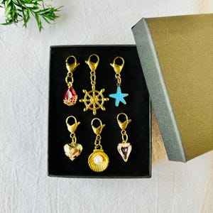May include: A collection of gold-toned charms with various designs, including a red teardrop, a ship's wheel, a blue starfish, a heart, a seashell with a pearl, and a pink teardrop. The charms are displayed in a black box with a gold lid.