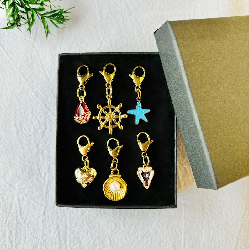 May include: A collection of gold-toned charms with various designs, including a red teardrop, a ship's wheel, a blue starfish, a heart, a seashell with a pearl, and a pink teardrop. The charms are displayed in a black box with a gold lid.