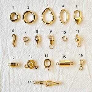 May include: A collection of gold-colored jewelry clasps and connectors, including spring rings, lobster clasps, heart-shaped clasps, and magnetic clasps. The clasps are displayed on a white, textured surface.