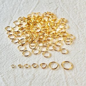 May include: A collection of gold-colored jump rings of various sizes, scattered on a white, textured surface. The rings are arranged in a pile, with a row of smaller rings displayed at the bottom. These jewelry-making supplies are ideal for crafting.