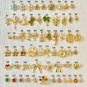 May include: A collection of gold-colored charms and pendants in various floral, leaf, and bow designs. Includes charms shaped like flowers, leaves, cacti, and bows. Some charms feature colorful accents, such as green, pink, and blue enamel.