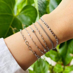 May include: A selection of silver bracelets in different styles. The bracelets feature heart-shaped links, beaded chains, and classic chain designs. The jewellery is shown on an arm, set against a background of green leaves.