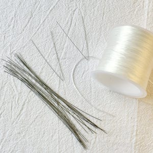 May include: Crafting supplies including a spool of clear elastic cord, a bundle of thin, dark-coloured wire stems, and several individual wire pieces. The items are arranged on a white, textured fabric surface.