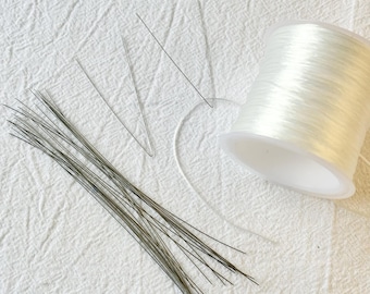 May include: A collection of craft supplies including a spool of clear elastic cord, a bundle of thin, dark-colored wire stems, and several individual wire pieces. The items are arranged on a white, textured fabric surface.