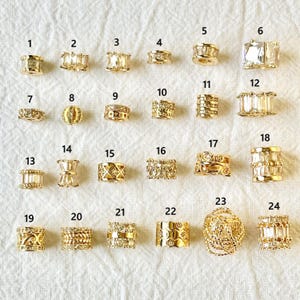 Può includere: Una collezione di 24 perline per gioielli color oro, ognuna con un design unico. Le perline variano nella forma, tra cui rettangolare, cilindrica e sferica, e sono decorate con accenti chiari e scintillanti. Le perline sono disposte su una superficie bianca.
