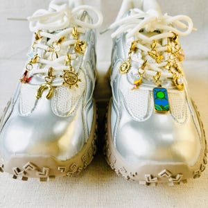 May include: A pair of silver sneakers with white laces and a beige sole. The sneakers are adorned with gold-colored charms, including a horseshoe, a ship's wheel, a sun, and a bird. The charms are attached to the laces.