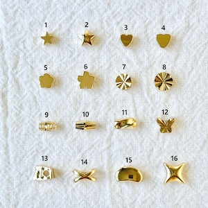 Könnte beinhalten: Eine Sammlung von sechzehn kleinen, goldfarbenen Charms in verschiedenen Formen, darunter Sterne, Herzen, Blumen und geometrische Designs. Die Charms werden auf einer weißen, strukturierten Oberfläche präsentiert.
