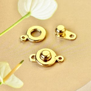 May include: Three gold-toned metal clasps for sewing or crafting projects. The clasps are round with a raised center button and a loop on each side.