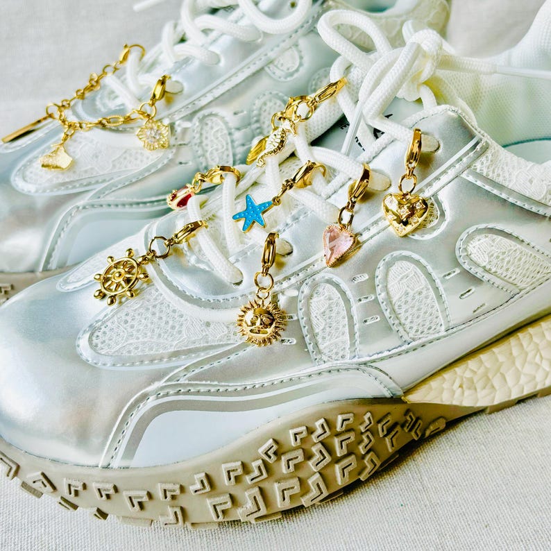 May include: Close-up of white sneakers with gold-colored charms. The charms include a sun, heart, and starfish. The sneakers have white laces and a silver iridescent finish. The soles are beige.