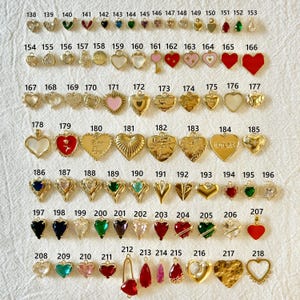 May include: A collection of gold-toned and colorful charms in various shapes, including hearts, teardrops, and geometric designs. Some charms feature gemstones, enamel, or text. The charms are arranged on a white surface.