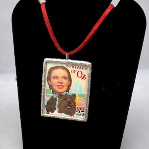 May include: A rectangular pendant featuring a vintage postage stamp design from "The Wizard of Oz." The stamp depicts a portrait of Dorothy and Toto, with the text "The Wizard of Oz" and "USA 25." The pendant is framed in silver and hangs from a red ribbon.