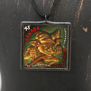 May include: Square pendant necklace featuring a vintage Canadian stamp with a goblin illustration. The stamp reads "CANADA" and "THE SUPERNATURAL GOBLIN". The pendant is framed in a dark metal and hangs from a black cord.