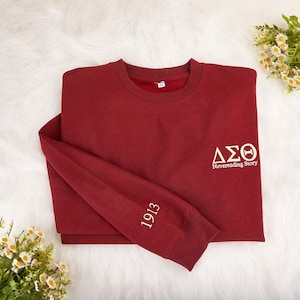 May include: A maroon sweatshirt with white text and graphics. The sleeve displays the number "1913" in white. The chest features Greek letters and the words "Neverending Story". The sweatshirt is folded on a white, fluffy surface.
