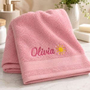 May include: A soft pink cotton bath towel featuring an embroidered design with the name Olivia in cursive script and a small yellow sun icon. The towel has a textured terry cloth surface and a decorative woven border detail along the bottom edge.