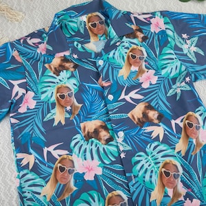 Könnte beinhalten: Ein kurzärmeliges Hemd mit tropischem Muster. Das Design zeigt blaue und grüne Blätter, rosa Blüten und Bilder einer Frau mit herzförmiger Sonnenbrille und einem Hundegesicht. Es hat einen klassischen Kragen und weiße Knöpfe.