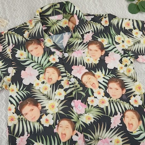 Op de afbeelding: Zwart overhemd met knopen met een tropisch bloemenpatroon met roze en gele bloemen en groene bladeren. Het shirt is versierd met meerdere afbeeldingen van een gezicht van een persoon.
