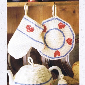 May include: A set of crocheted kitchen accessories in white with red and blue accents. The set includes a teapot cozy, a pot holder, an oven mitt, and an egg cozy. Each piece features a heart design.