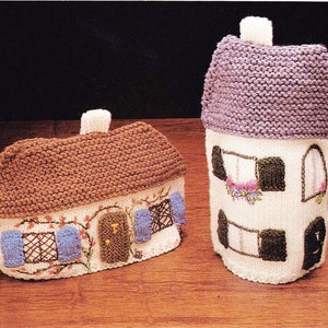 Könnte beinhalten: Zwei gestrickte Häuser, eines mit einem braunen Dach und das andere mit einem violetten Dach. Beide Häuser haben weiße Wände mit gestickten Details, darunter Fenster, Türen und Blumen.