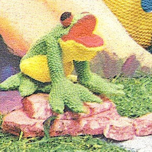 May include: A green crocheted frog with a yellow belly and a red mouth sitting on a pink rock. The frog is sitting on a bed of green grass.
