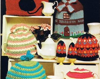 May include: A collection of knitted teapot cosies in various colours and designs, including a cat, a windmill, stripes, and a flower pattern. The cosies are displayed on a shelf with teapots and other tea accessories.