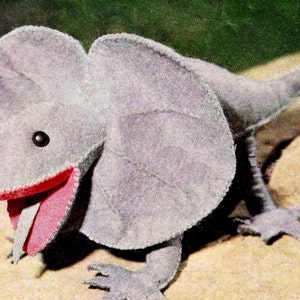 Peut inclure: Un lézard en feutre gris avec un grand cou à volants et une bouche rose ouverte. Le lézard a des yeux noirs et est assis sur une surface beige.